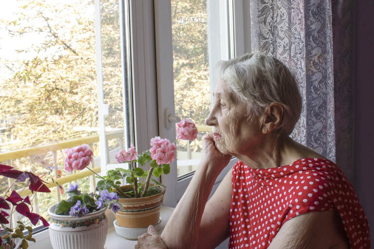 Old Lonely Woman Sitting Near The Window In His House With Flowe